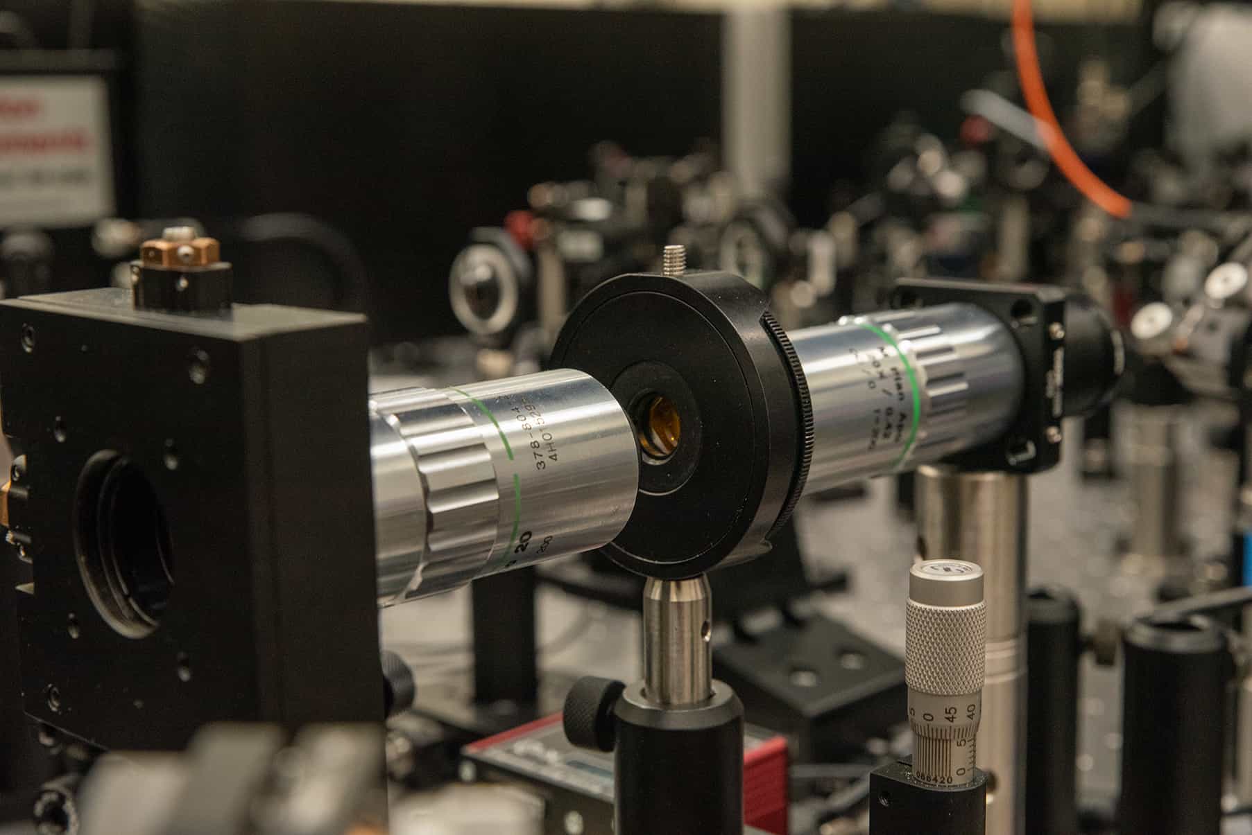Microscope objectives and lens mounts lined up along a laser beam path on an optical table, with more optics blurred in the background.