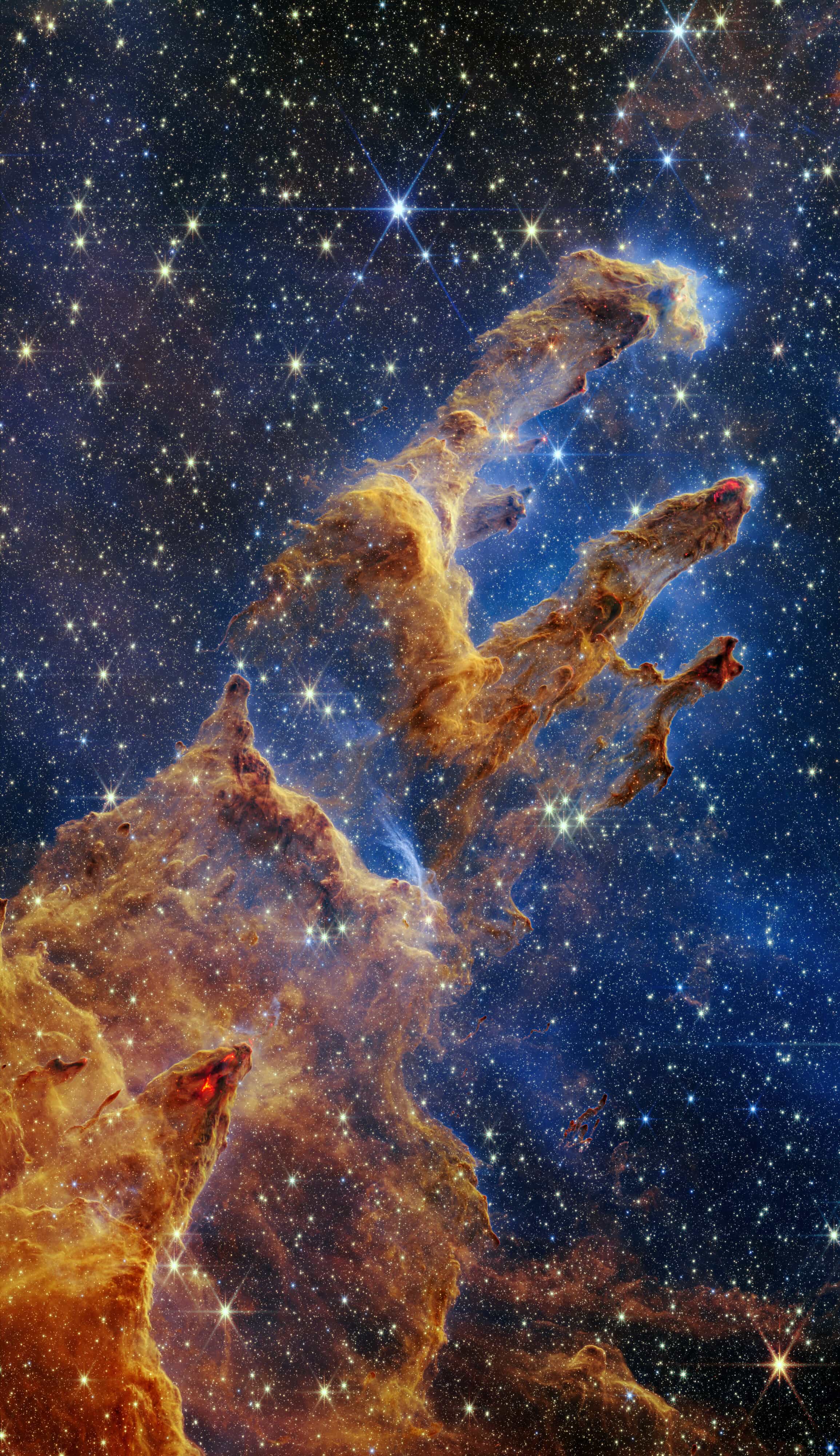 Webb near-infrared view of the Pillars of Creation in the Eagle Nebula: tall columns of orange-brown gas and dust rising through a blue haze crowded with stars.
