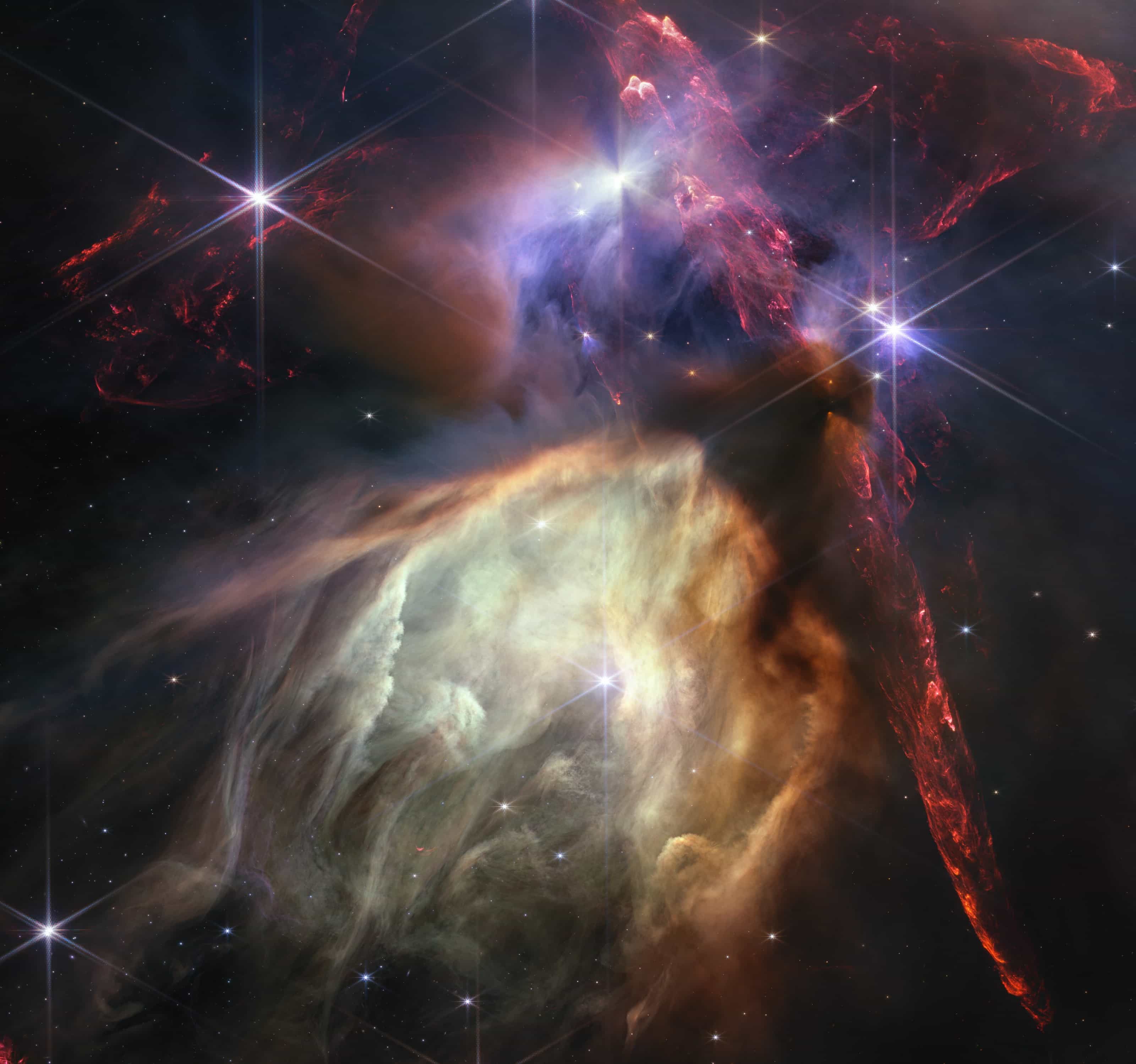 Webb image of the Rho Ophiuchi star-forming cloud: glowing cream and brown gas lit by young stars, streaked with red jets, with bright stars showing Webb's six-pointed diffraction spikes.