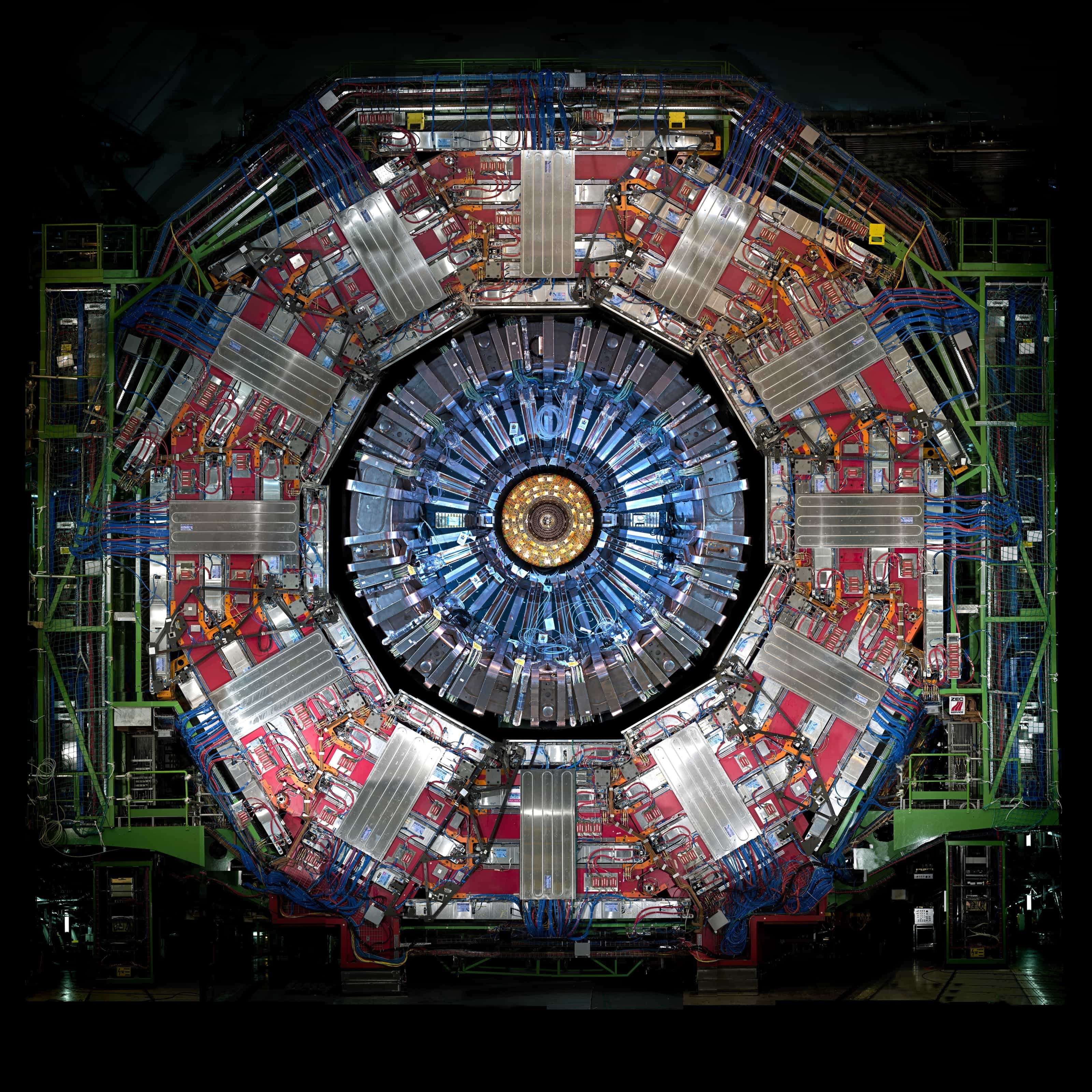 Head-on view of the open CMS detector at CERN: a twelve-sided ring of red and silver muon chambers and cabling surrounds a glowing blue-and-gold core, against darkness.