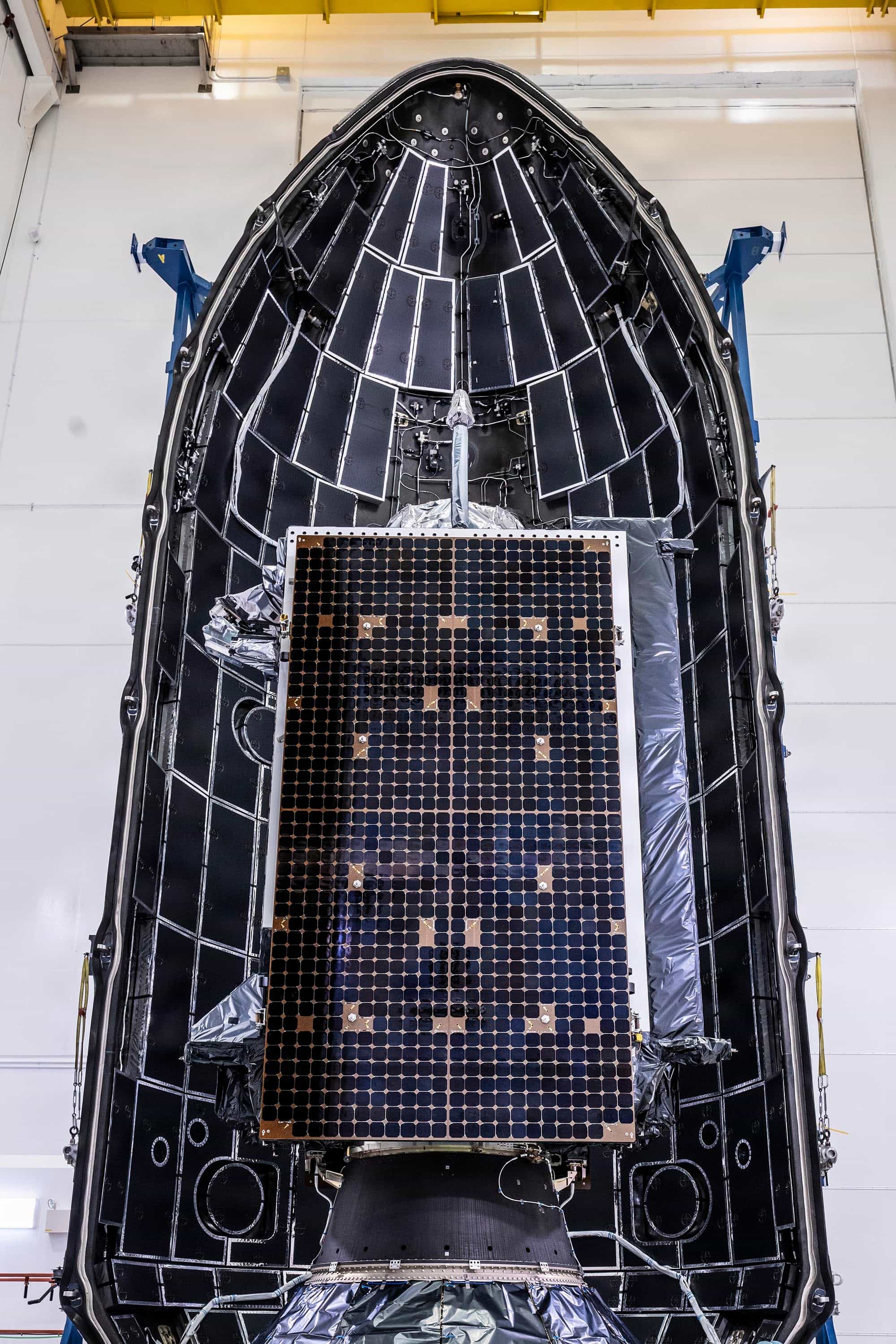 A GPS III satellite, its dark solar-panel wing facing the camera, sits inside one half of a SpaceX Falcon 9 payload fairing in a white processing hall before the GPS III-8 launch, April 2026.
