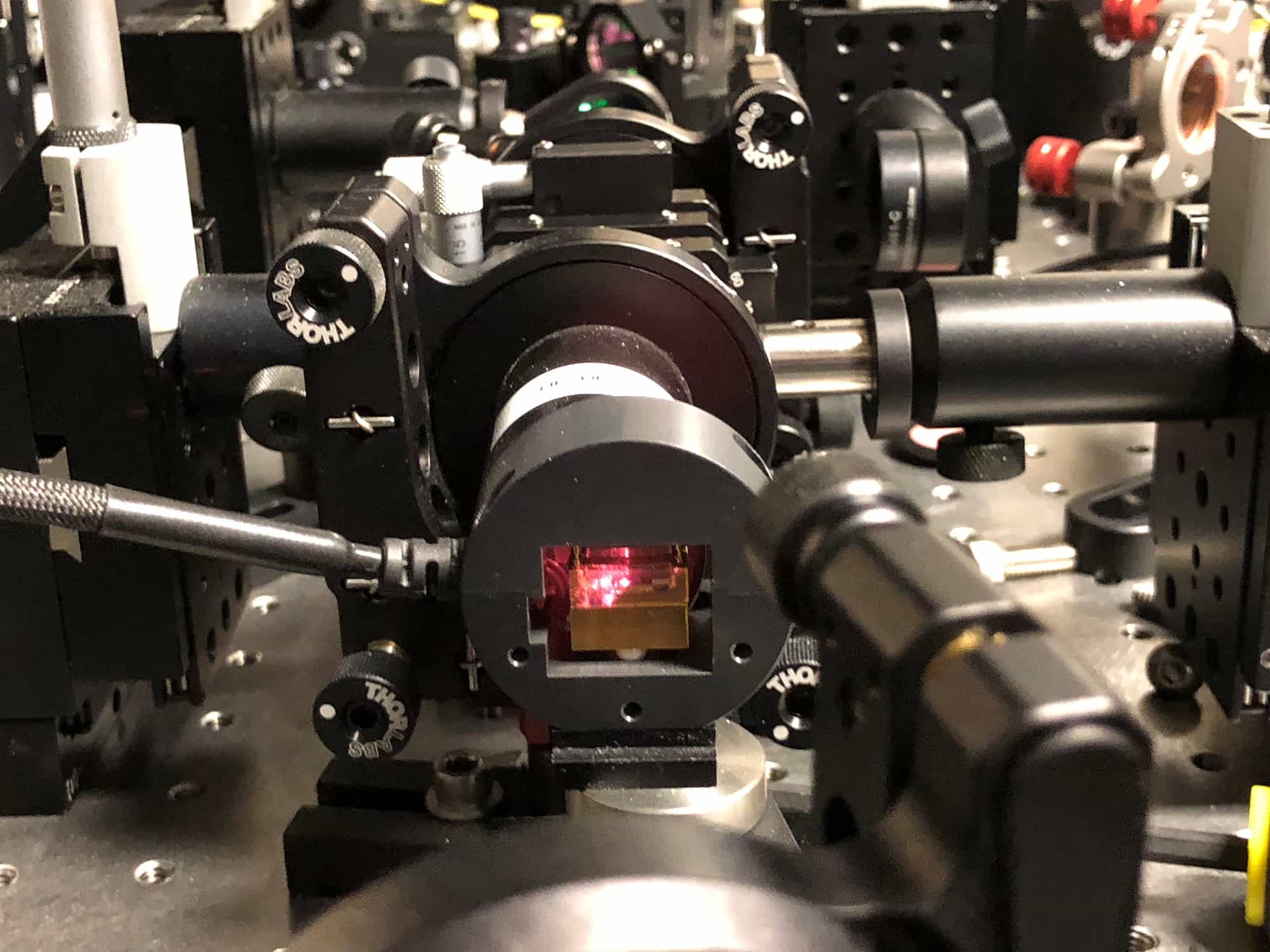 Close-up of black optical mounts on a lab bench, with a red glow at the centre from the crystal that converts laser light into pairs of photons.
