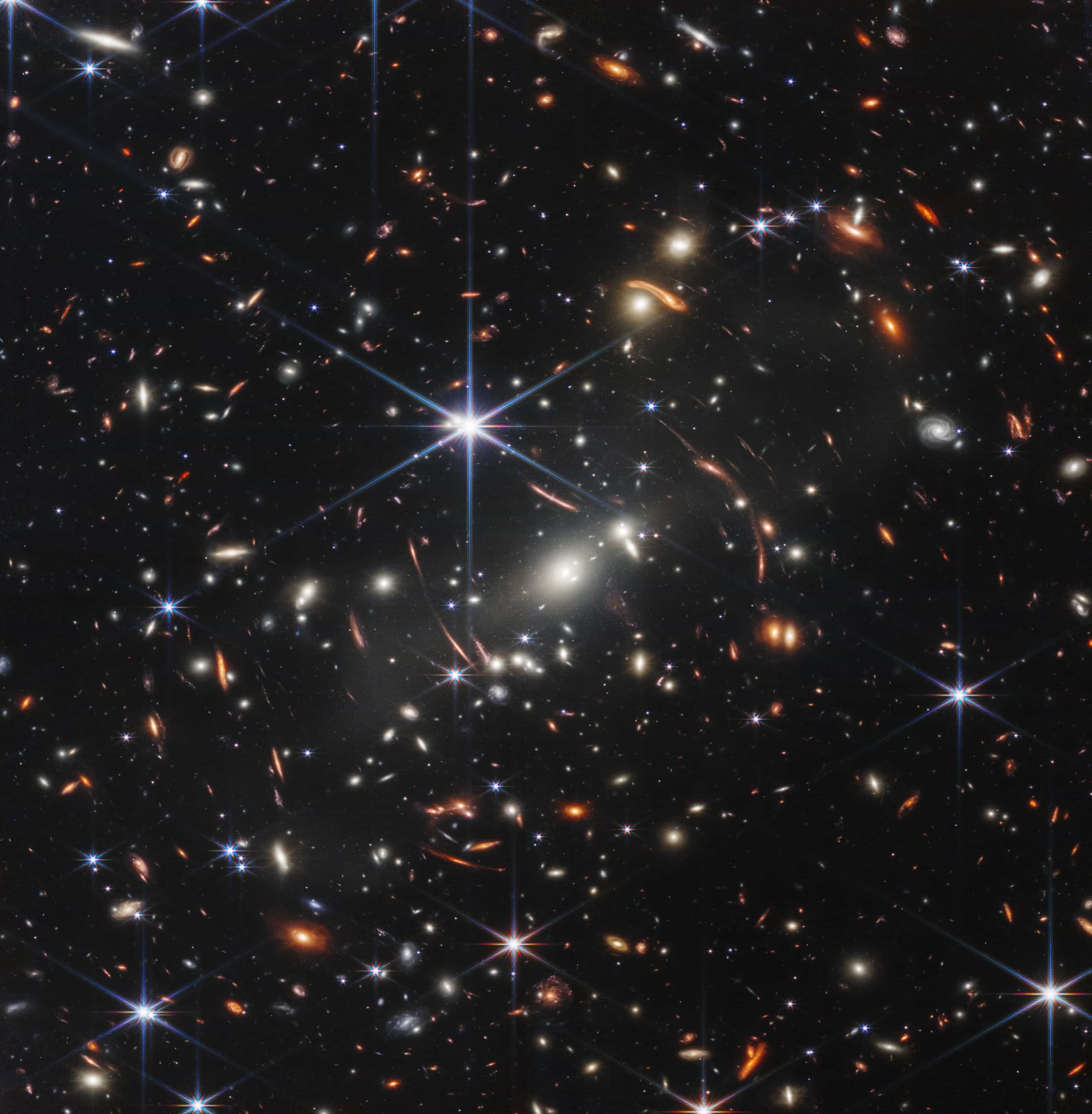 Webb's first deep field, galaxy cluster SMACS 0723: hundreds of galaxies on black, many distant ones stretched into thin orange arcs by the cluster's gravity, plus a few spiky foreground stars.