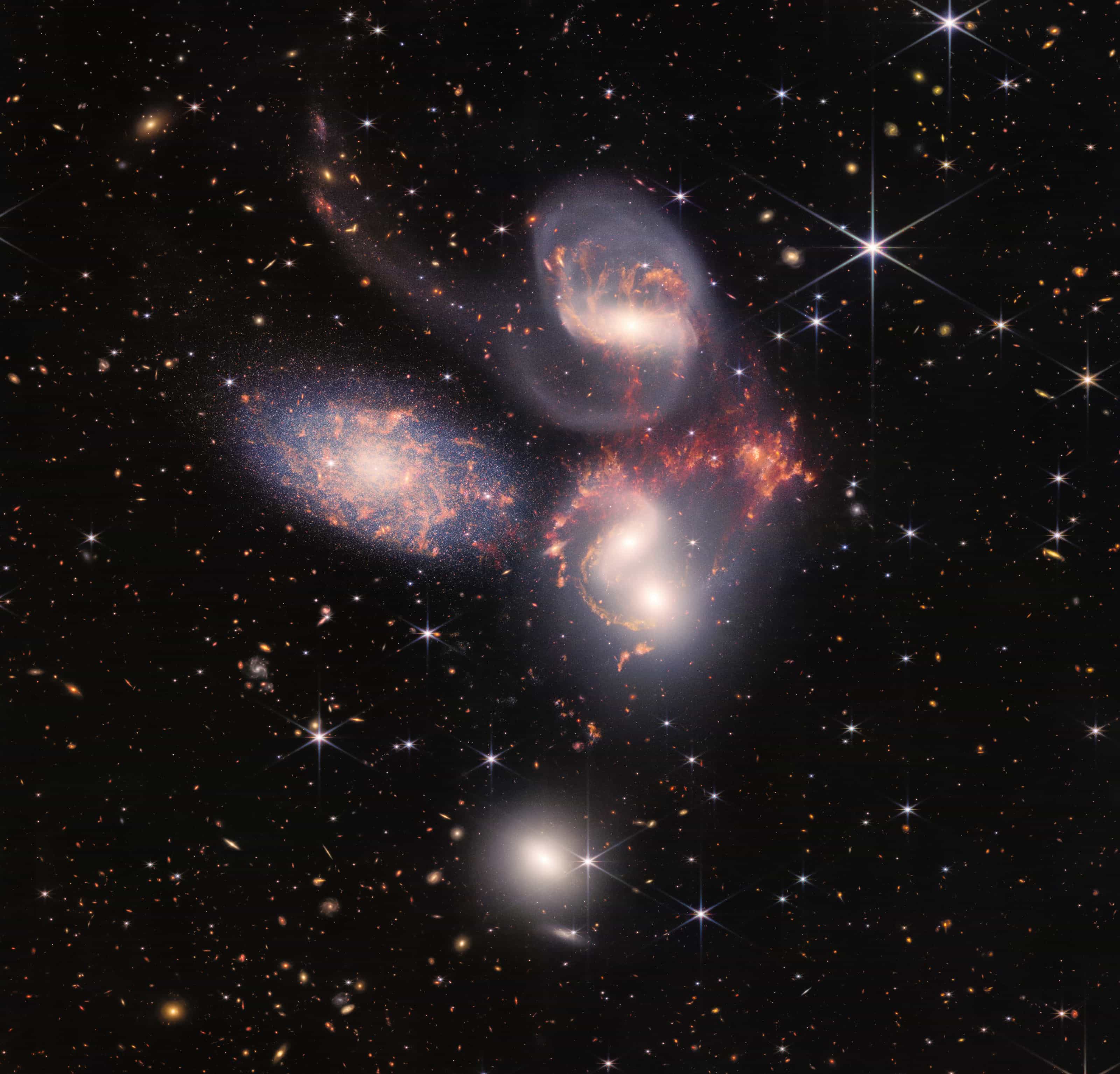 Webb image of Stephan's Quintet: five galaxies, four of them interacting, with tails of gas and red-orange star-forming regions, against a black field of distant galaxies.