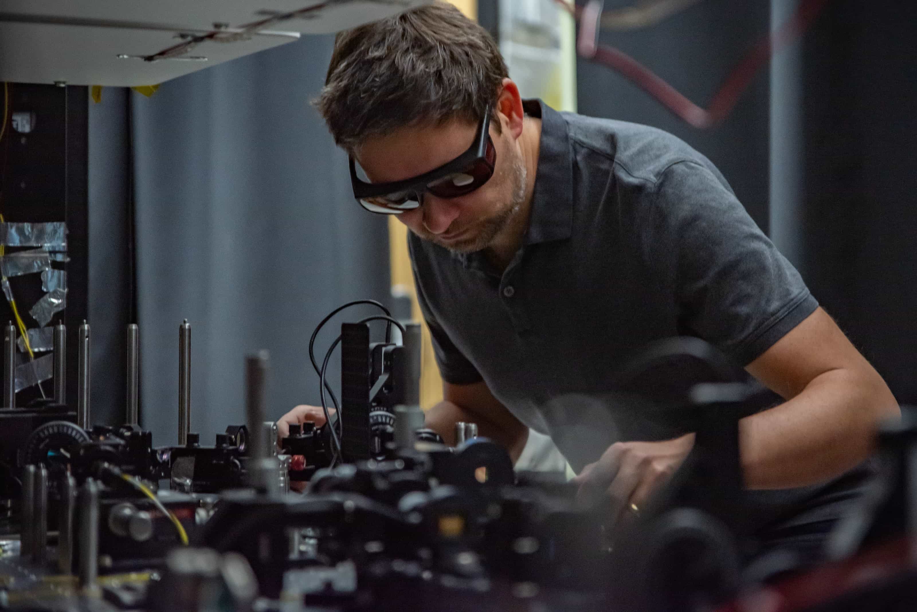 NIST physicist Krister Shalm, wearing laser-safety glasses, adjusts components on a crowded optical table in a dark laboratory.