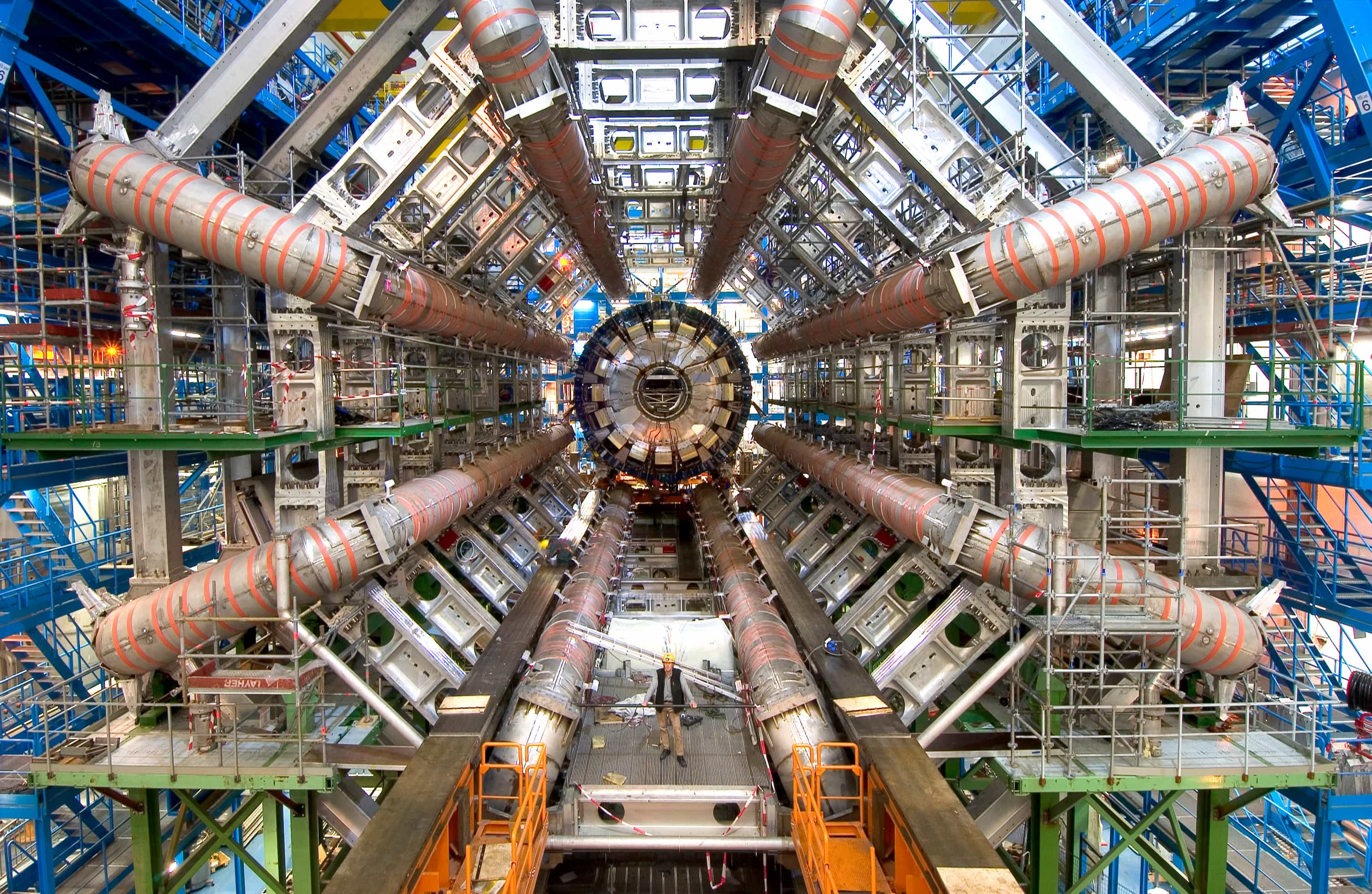The ATLAS detector cavern at CERN in 2005: eight huge silver toroid magnet tubes radiate around the cylindrical calorimeter at the centre, with a person standing on the floor below for scale.
